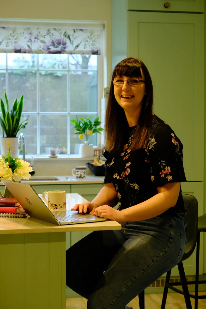 Copywriter for small businesses and founders michelle works at her laptop in a green kitchen, she has dark hair with a full fringe and glasses